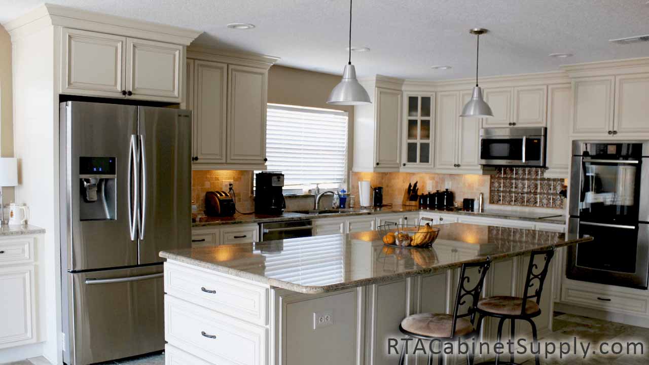 Country Cream kitchen angle view with an island, a glass door cabinet, wall and base cabinets, lighting and a fridge.